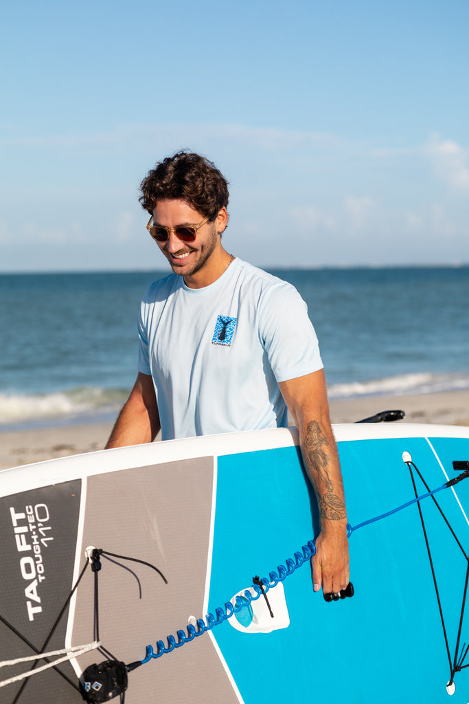 Man wearing Tunaskin Coastal Collective shirt on beach with paddleboard