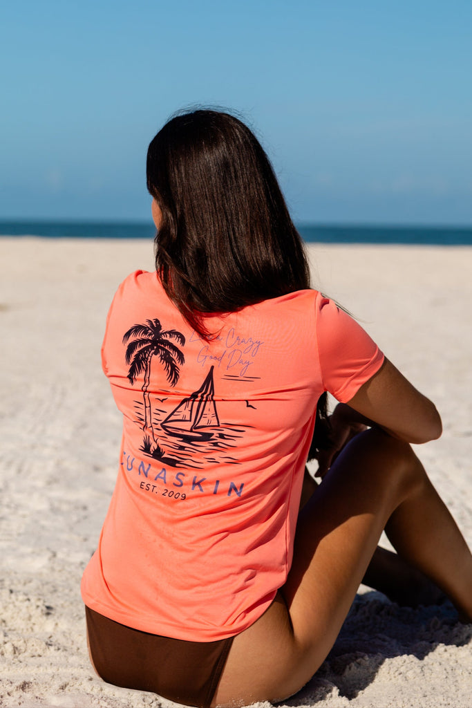 Woman sitting on a sandy beach wearing a coral t-shirt with a palm tree and sailboat design.