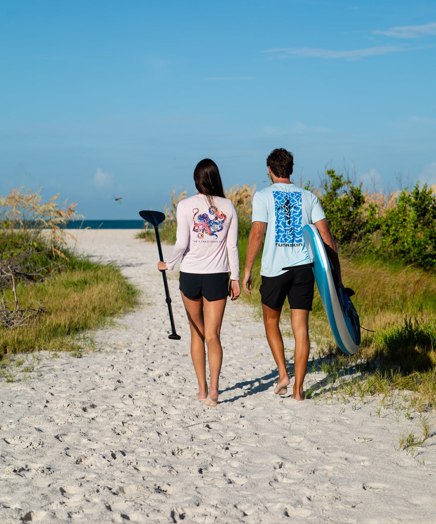 Two people wearing tunaskin shirts walking on a sandy path with paddle board