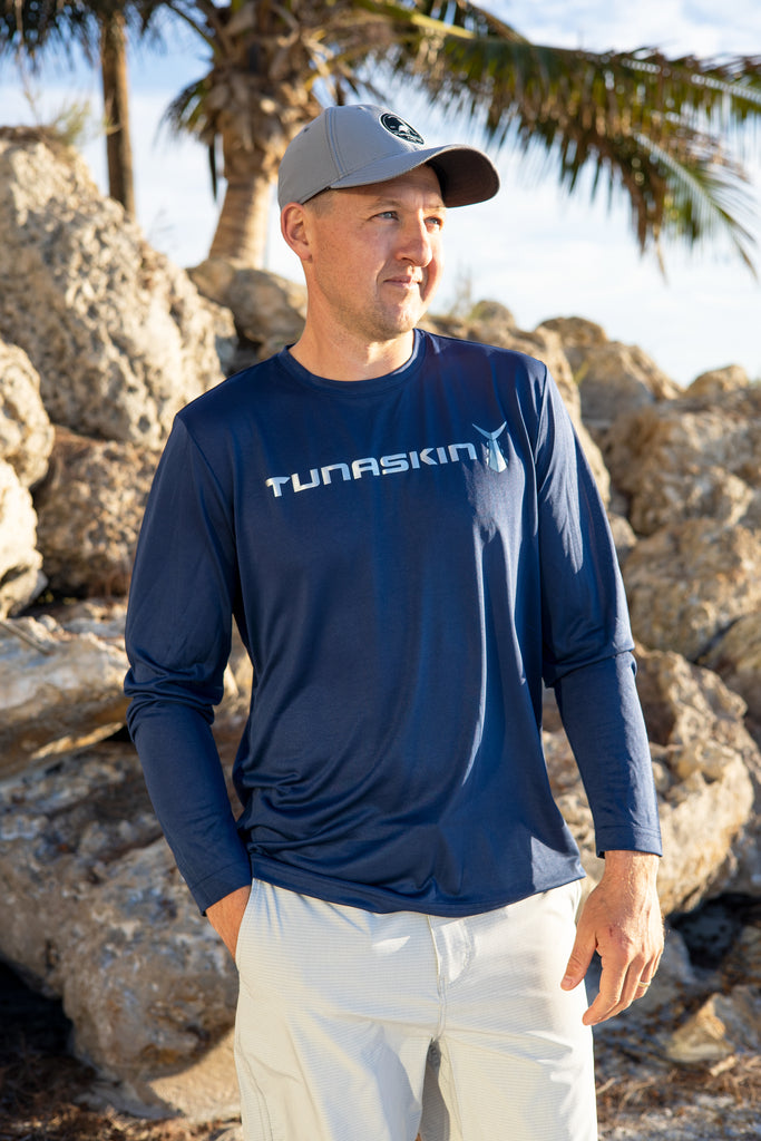 Man wearing a navy blue shirt with 'Tunaskin' logo, standing in front of rocky terrain and palm trees.