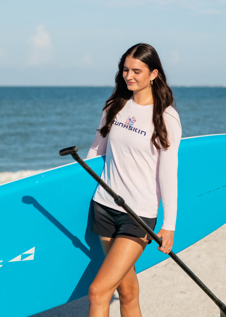 Woman holding a paddle and a blue paddleboard on a beach with tunaskin long sleeve