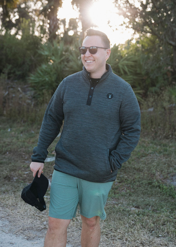 Man wearing sunglasses and a dark green pullover with shorts, standing outdoors.