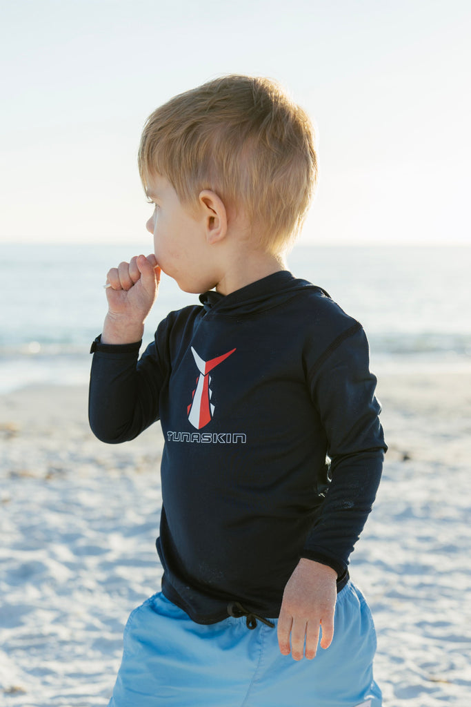 Child in navy Tunaskin hoodie and blue shorts standing on a sandy beach.